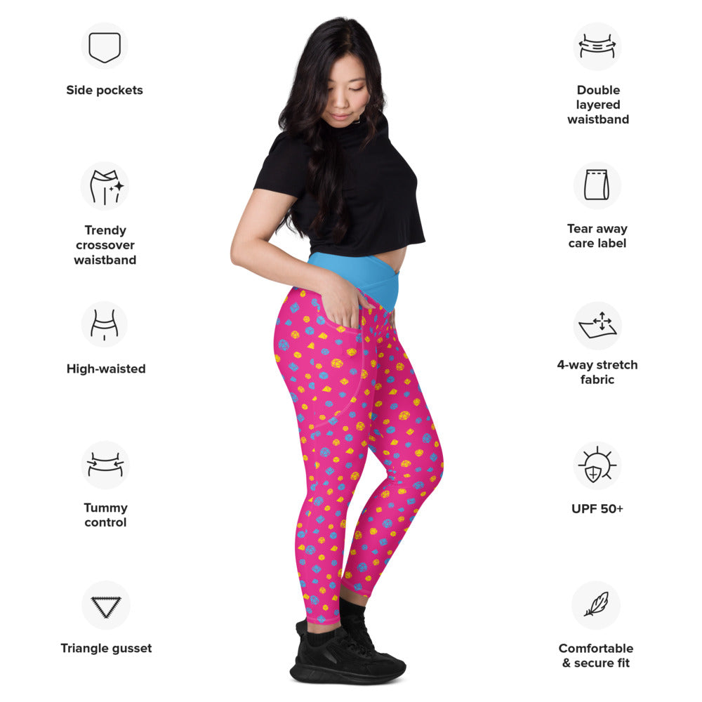 Light-skinned dark-haired female-presenting model wearing pansexual dice leggings. she is facing right and has a hand in the pocket. she is surrounded by product specs: "side pockets, trendy crossover waistband, high-waisted, tummy control, triangle gusset, double layered waistband, tear away care label, 4-way stretch fabric, UPF 50+, comfortable & secure fi
