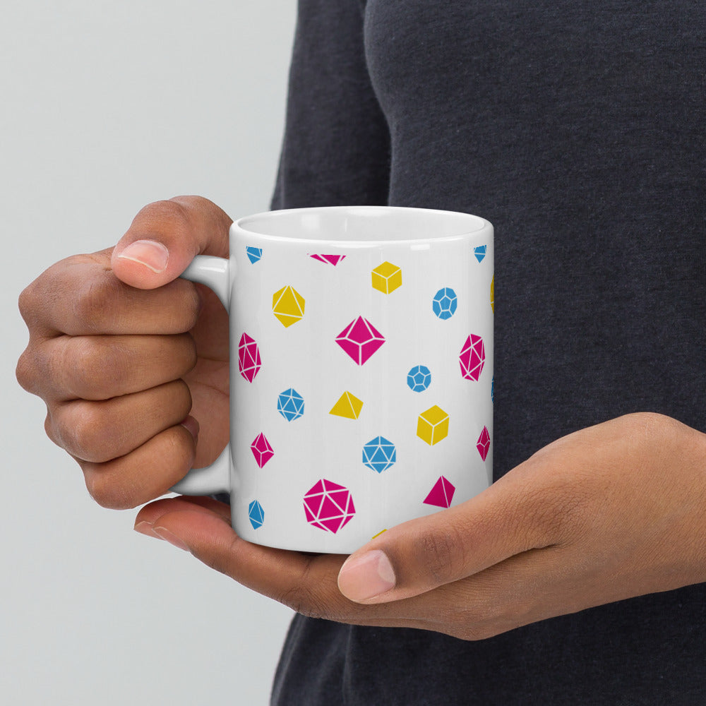 white mug being held by the handle by a dark-skinned set of hands. It has an all-over print of polyhedral d&d dice in the pansexual colors of pink, blue, and yellow