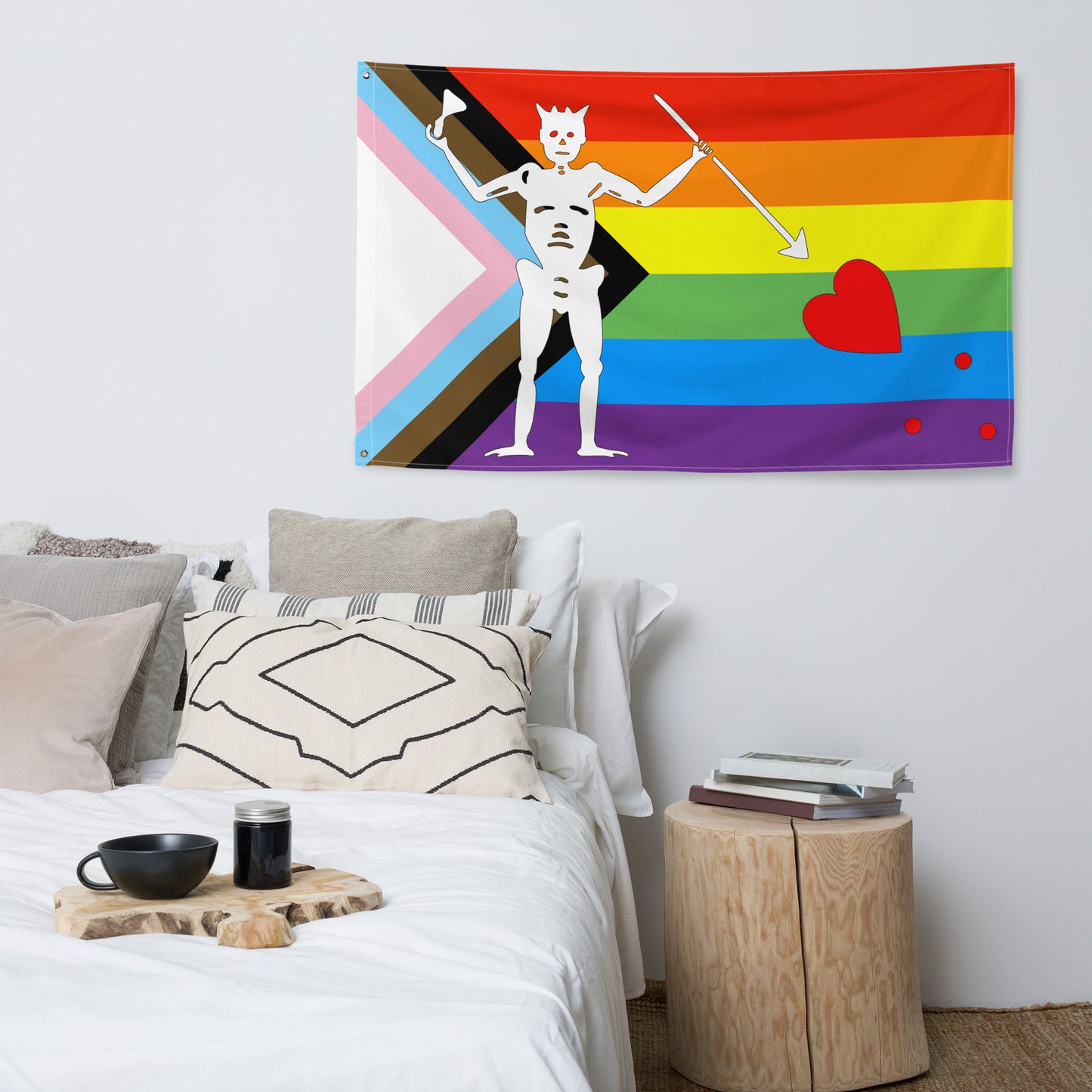 the progress pride blackbeard pride flag hanging on a white wall above a bed and a side table