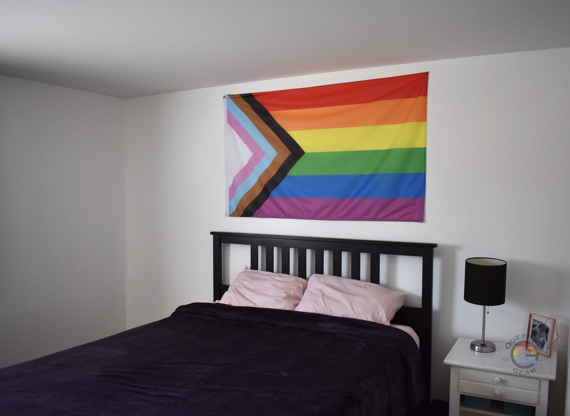 3’x5’ progress pride flag hanging horizontally on the wall of a bedroom centered above a bed with a purple blanket