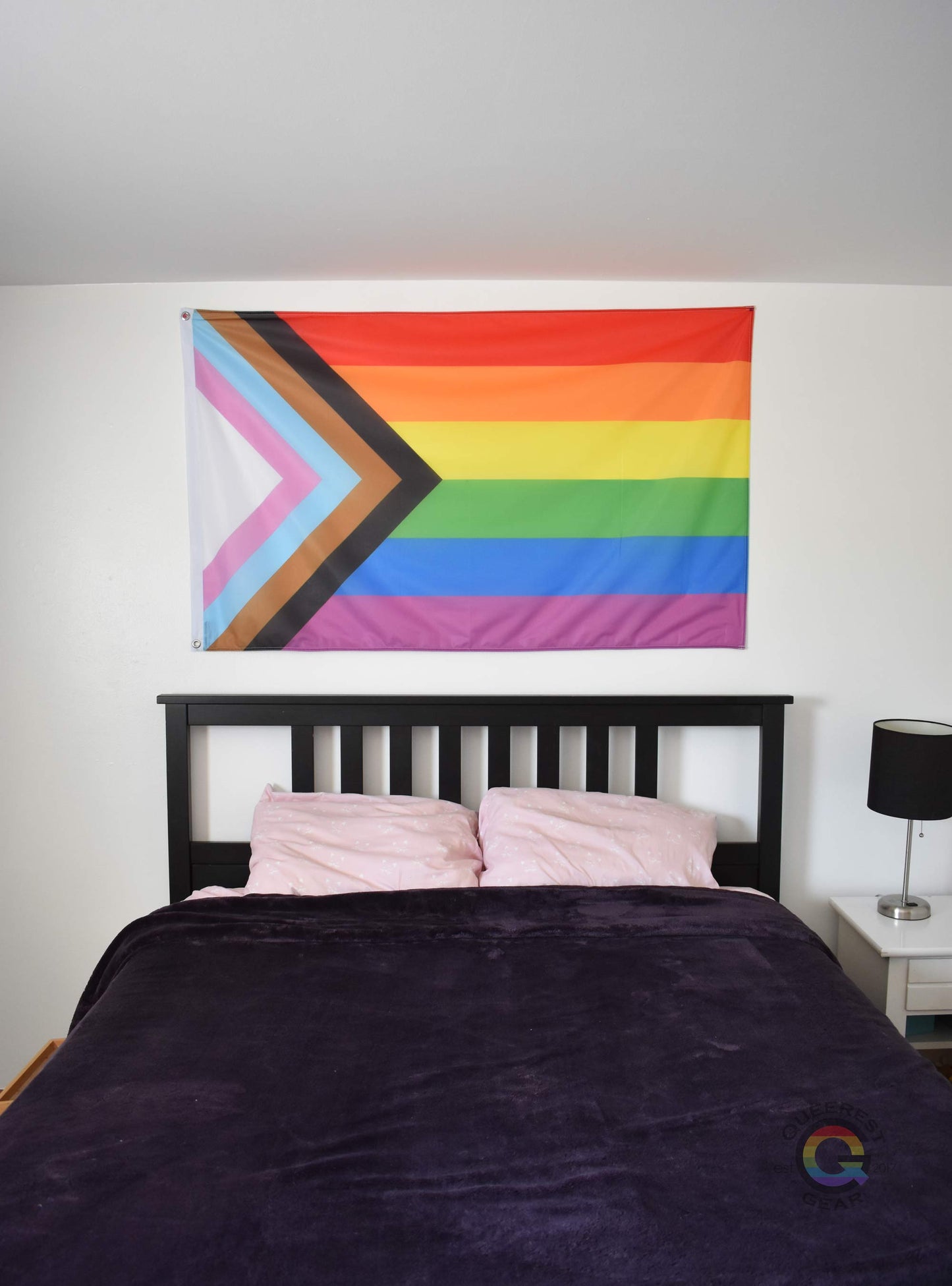 3’x5’ progress pride flag hanging horizontally on the wall of a bedroom centered above a bed with a purple blanket
