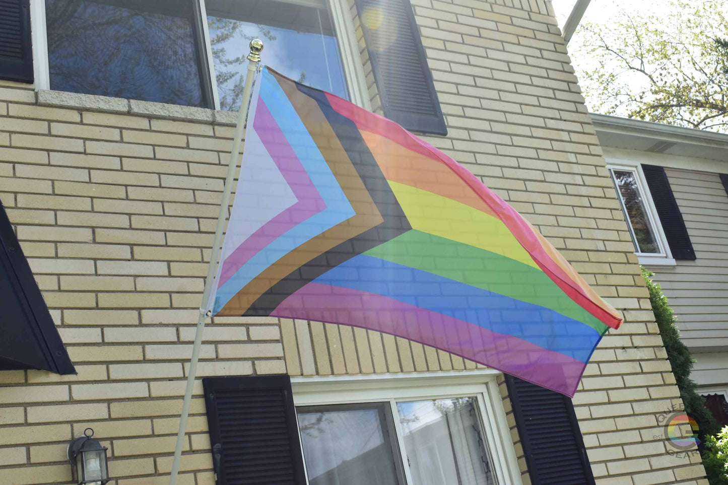 3’x5’ progress pride flag hanging from a flagpole on the outside of a light brick house with dark shutters