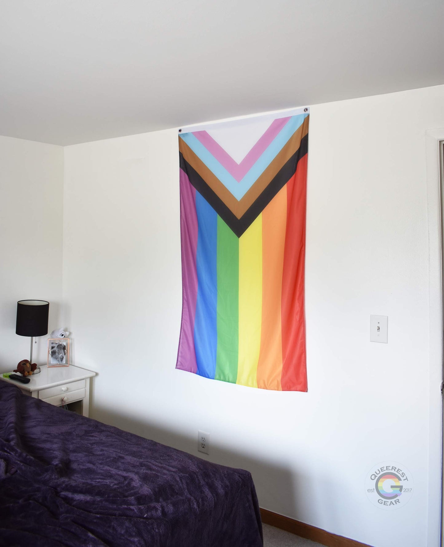  3’x5’ progress flag hanging vertically on the wall of a bedroom with a nightstand and a bed