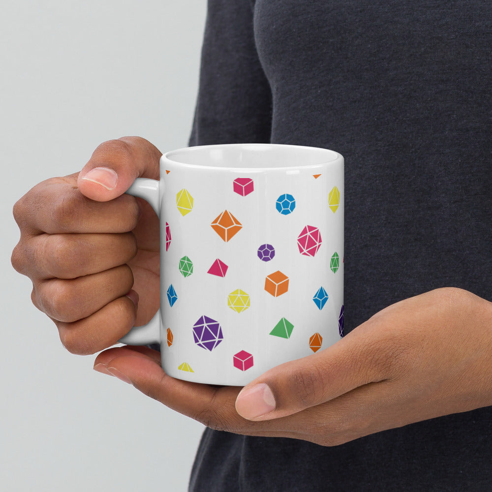 white mug being held by the handle by a dark-skinned set of hands. It has an all-over print of polyhedral d&d dice in rainbow colors