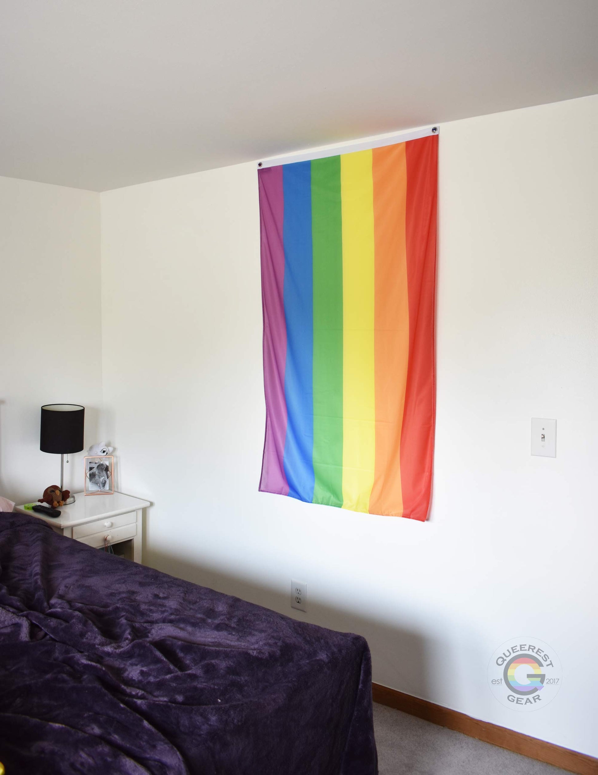  3’x5’ rainbow flag hanging vertically on the wall of a bedroom with a nightstand and a bed