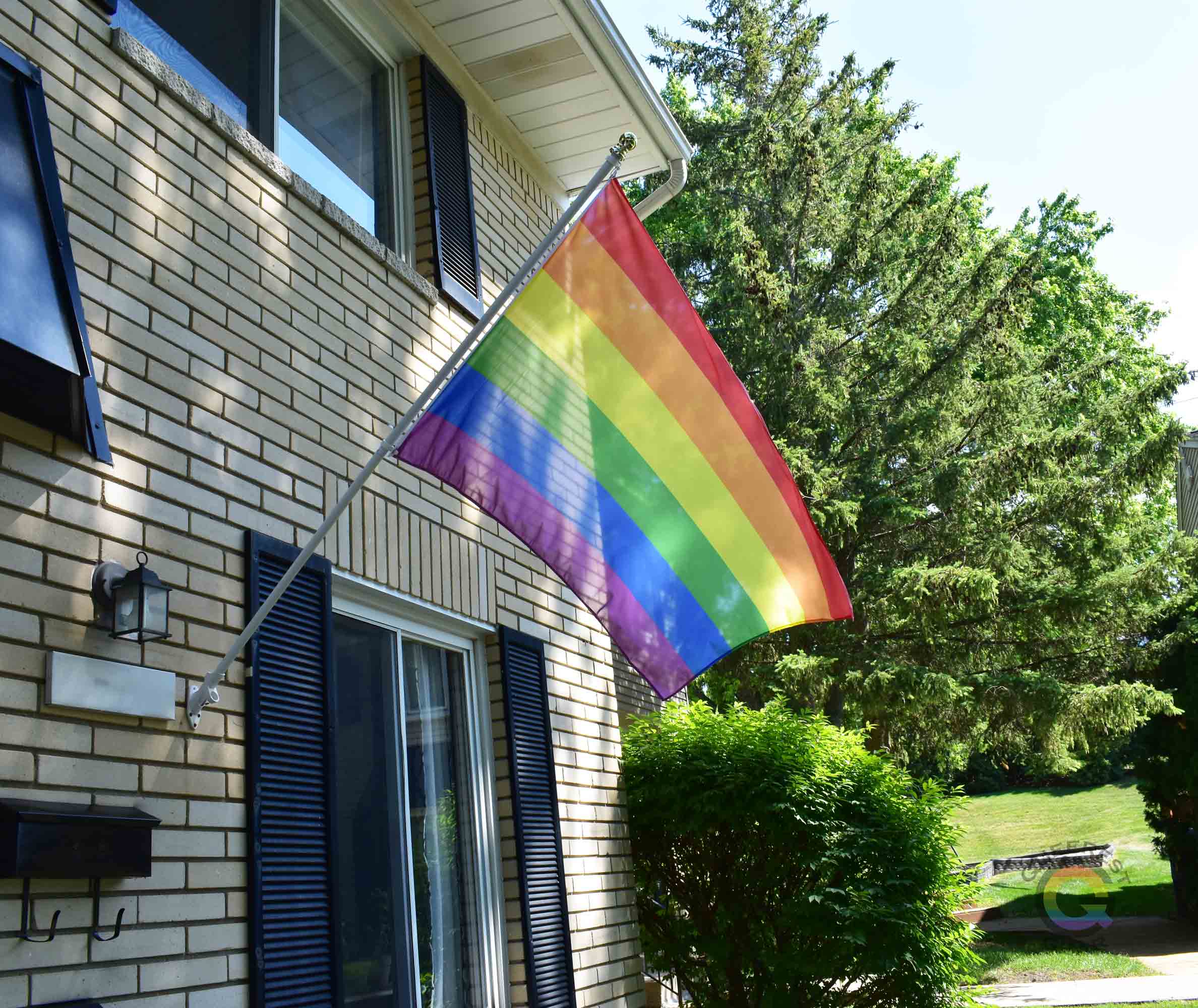 3’x5’ rainbow pride flag hanging from a flagpole on the outside of a light brick house with dark shutters
