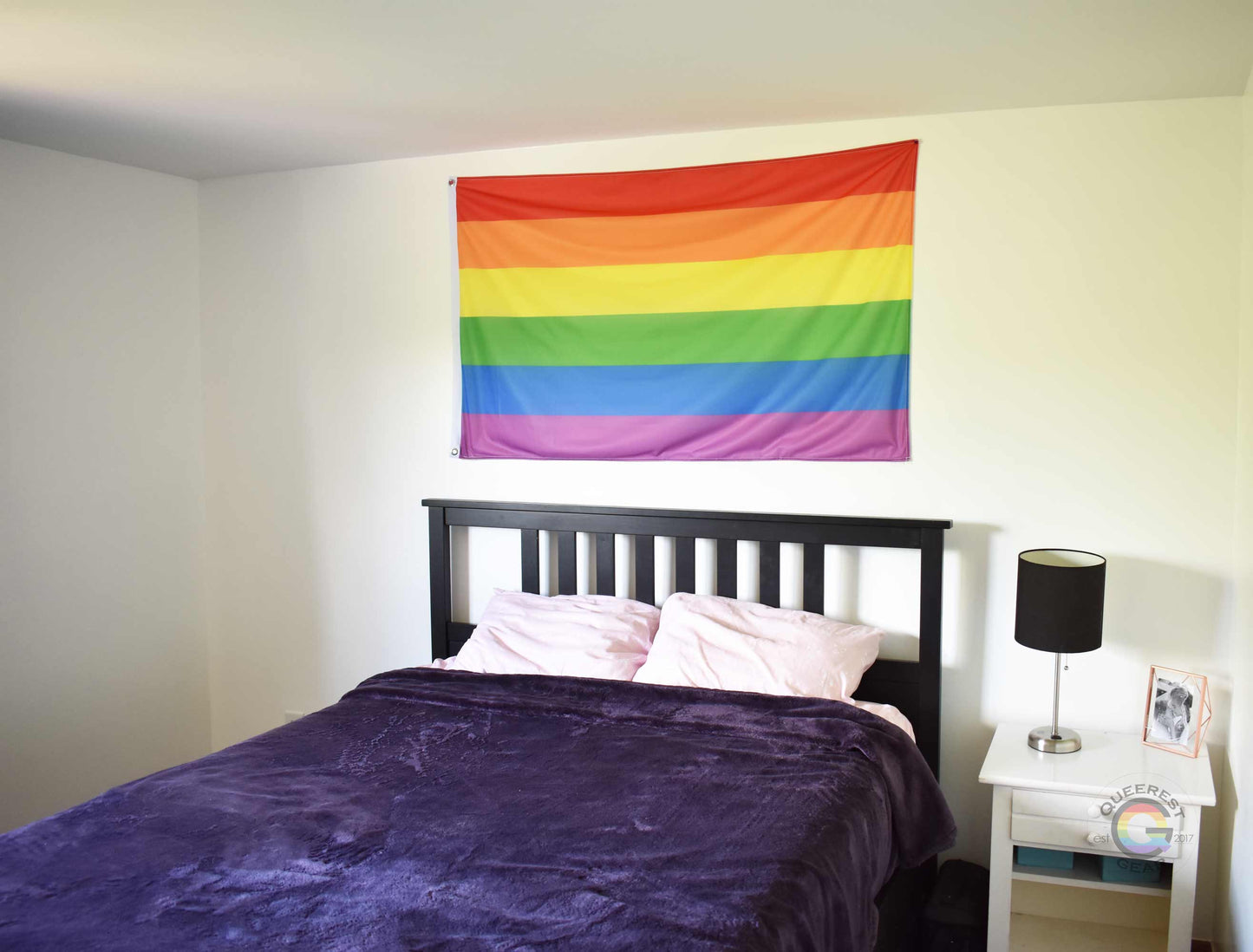3’x5’ rainbow pride flag hanging horizontally on the wall of a bedroom centered above a bed with a purple blanket