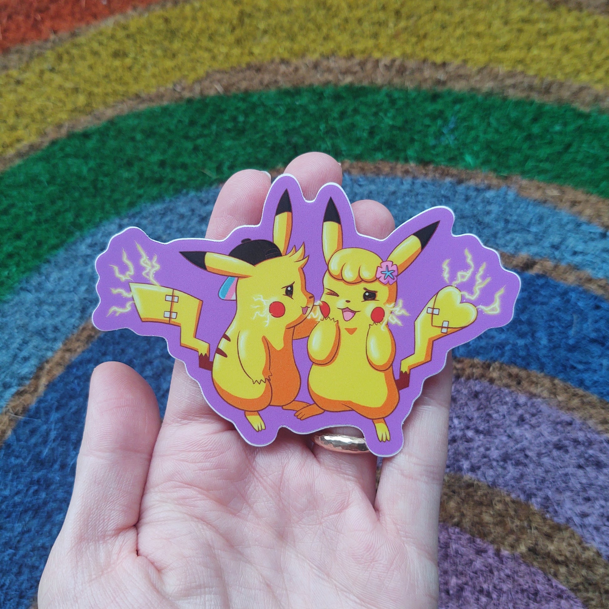 the trans for trans sticker held by a light-skinned hand on a rough rainbow background. the sticker is a pair of transgender animals with electric current flowing and a purple outline