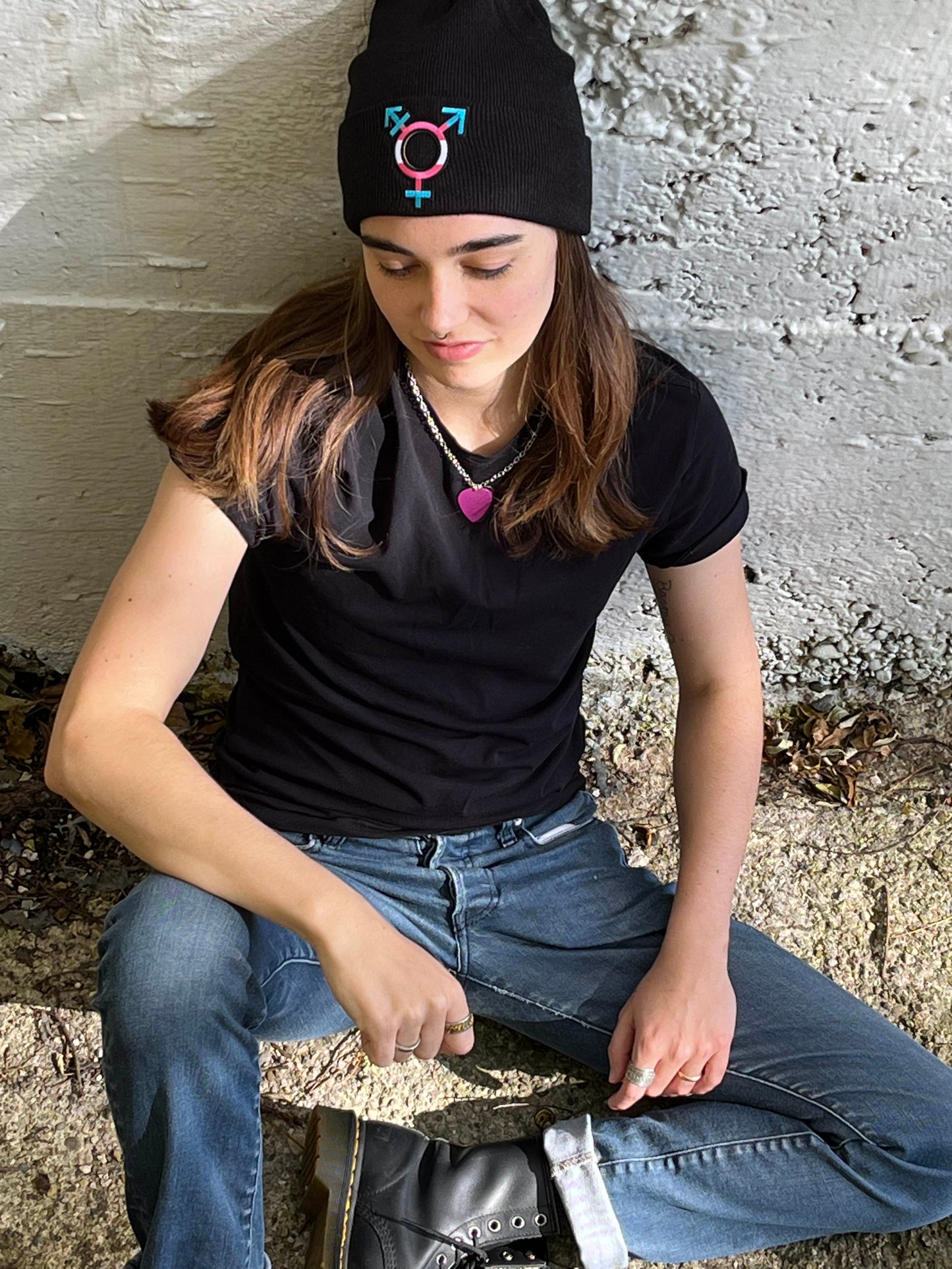 person sitting against white concrete wall looking down while wearing transgender pride beanie