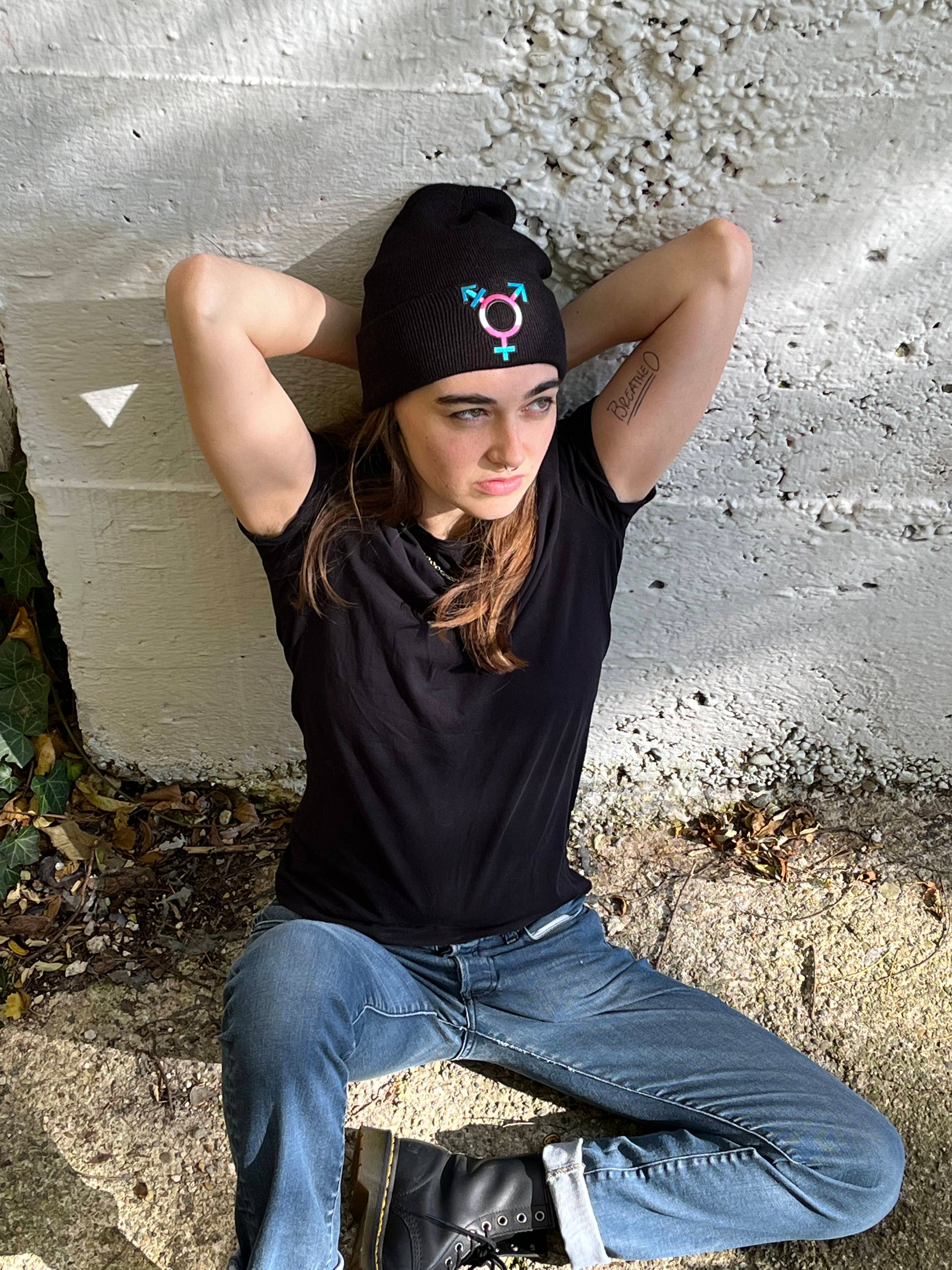 person sitting leaning against white concrete with hands behind their head wearing the transgender beanie