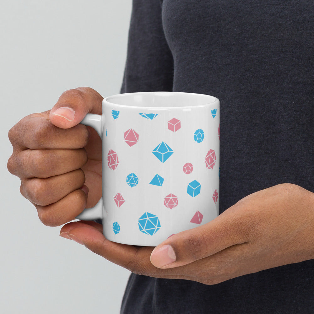 white mug being held by the handle by a dark-skinned set of hands. It has an all-over print of polyhedral d&d dice in the transgender colors of pink and blue