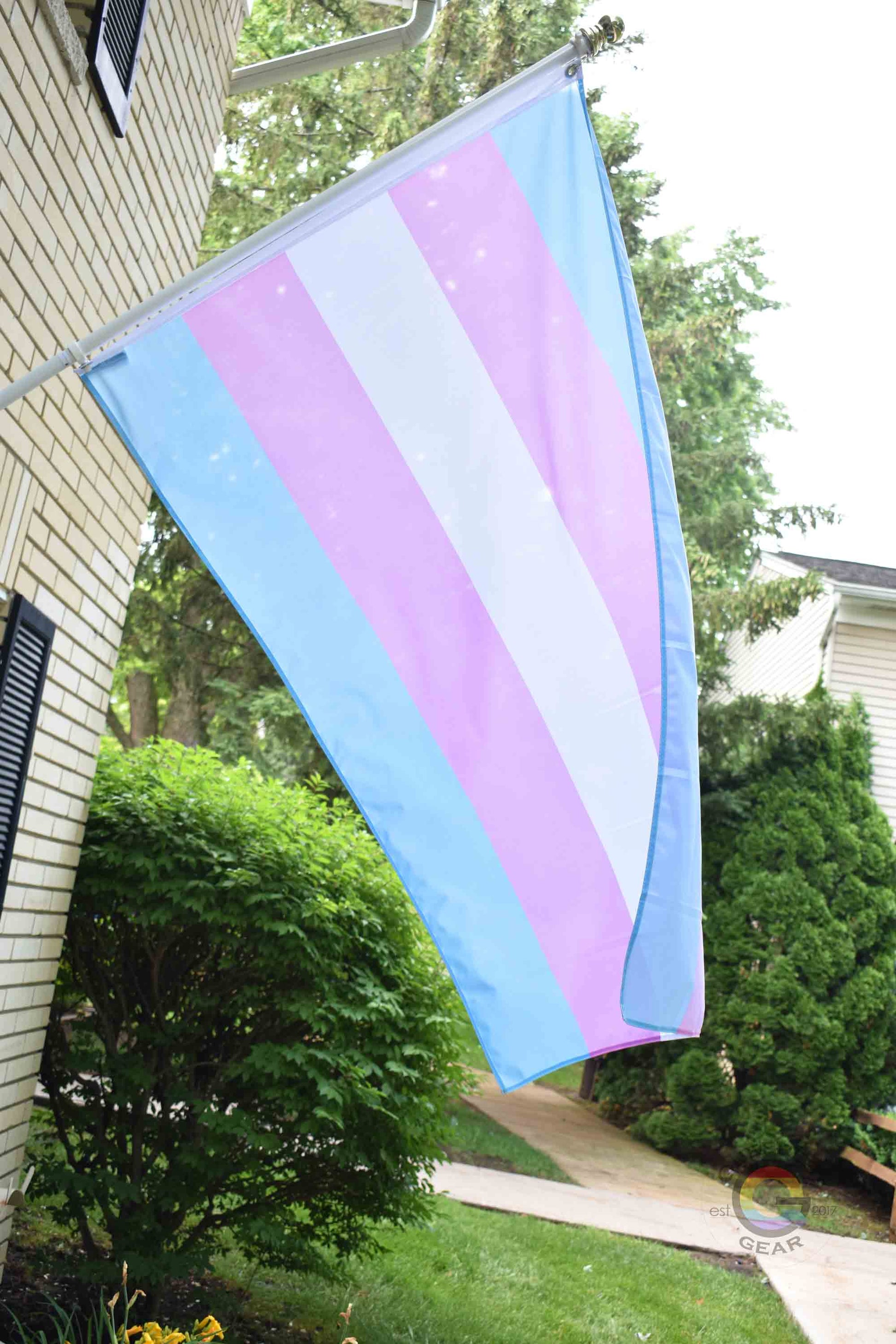 3’x5’ transgender pride flag hanging from a flagpole on the outside of a light brick house with dark shutters