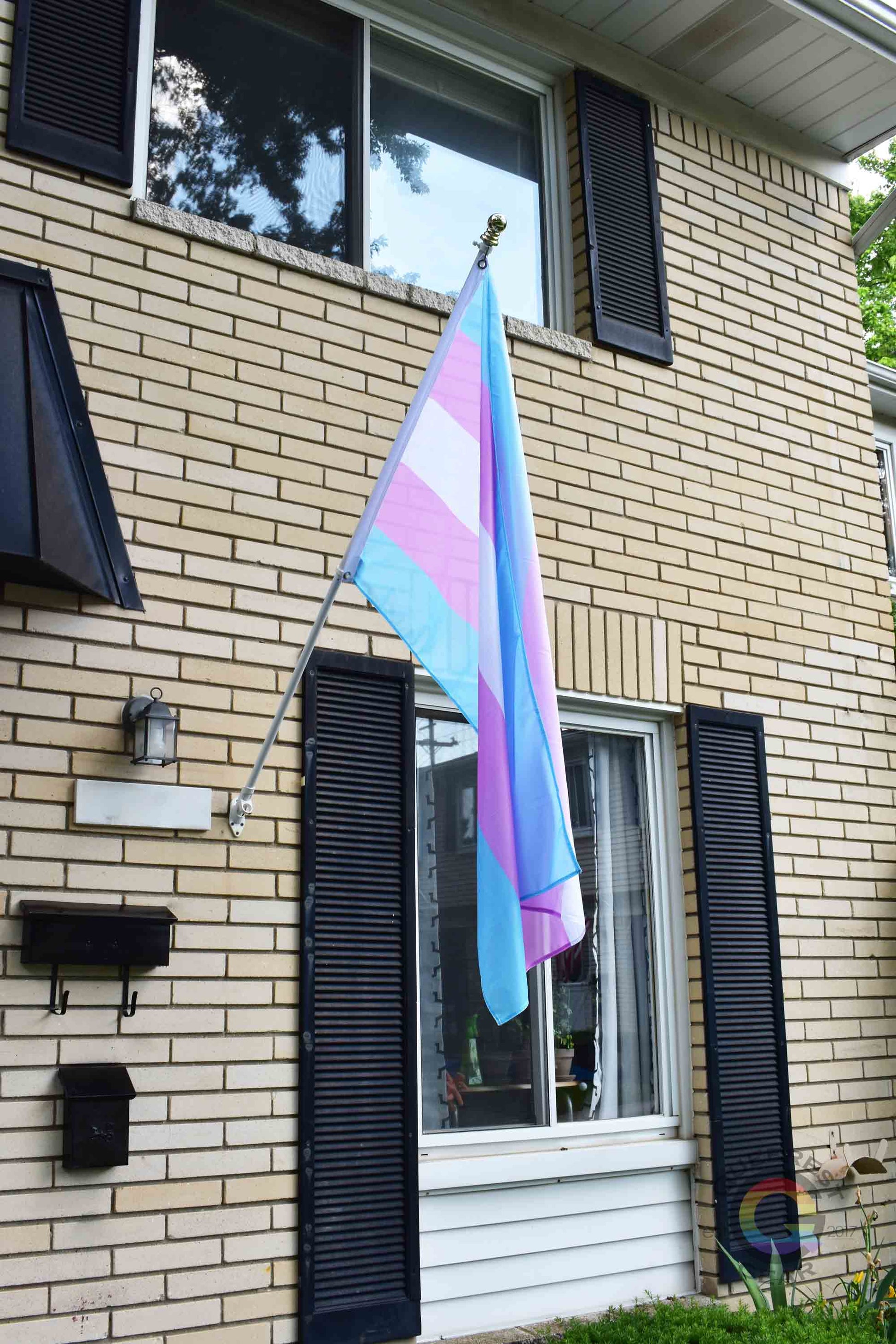 3’x5’ transgender pride flag hanging from a flagpole on the outside of a light brick house with dark shutters